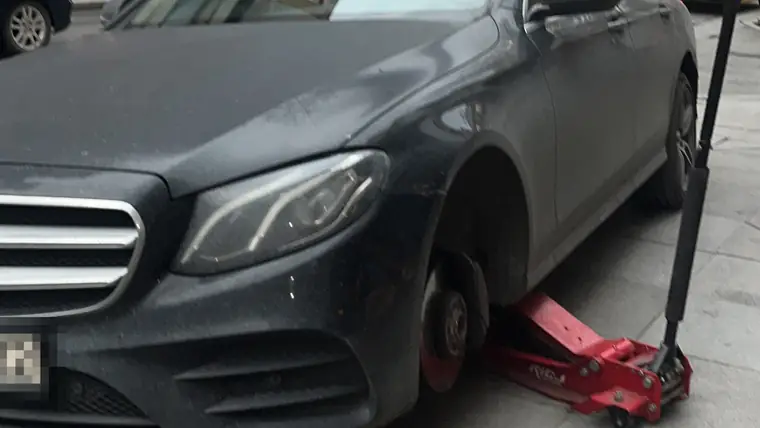 Şehirde kaldırım kenarında bir otomobilin ön tekerleği sökülmüş; araç kırmızı krikoda, fren diski açıkta görünüyor.