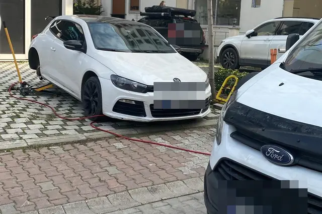 Apartman önündeki parke taşlı alanda krikoya alınmış beyaz bir otomobil; yerde kırmızı hava hortumu, önde beyaz bir hafif ticari araç.