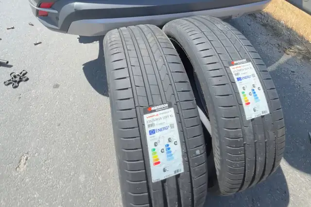 Asfaltta bir aracın arkasında, etiketleri üzerinde duran iki yeni lastik; yanda sökülmüş bijonlar.